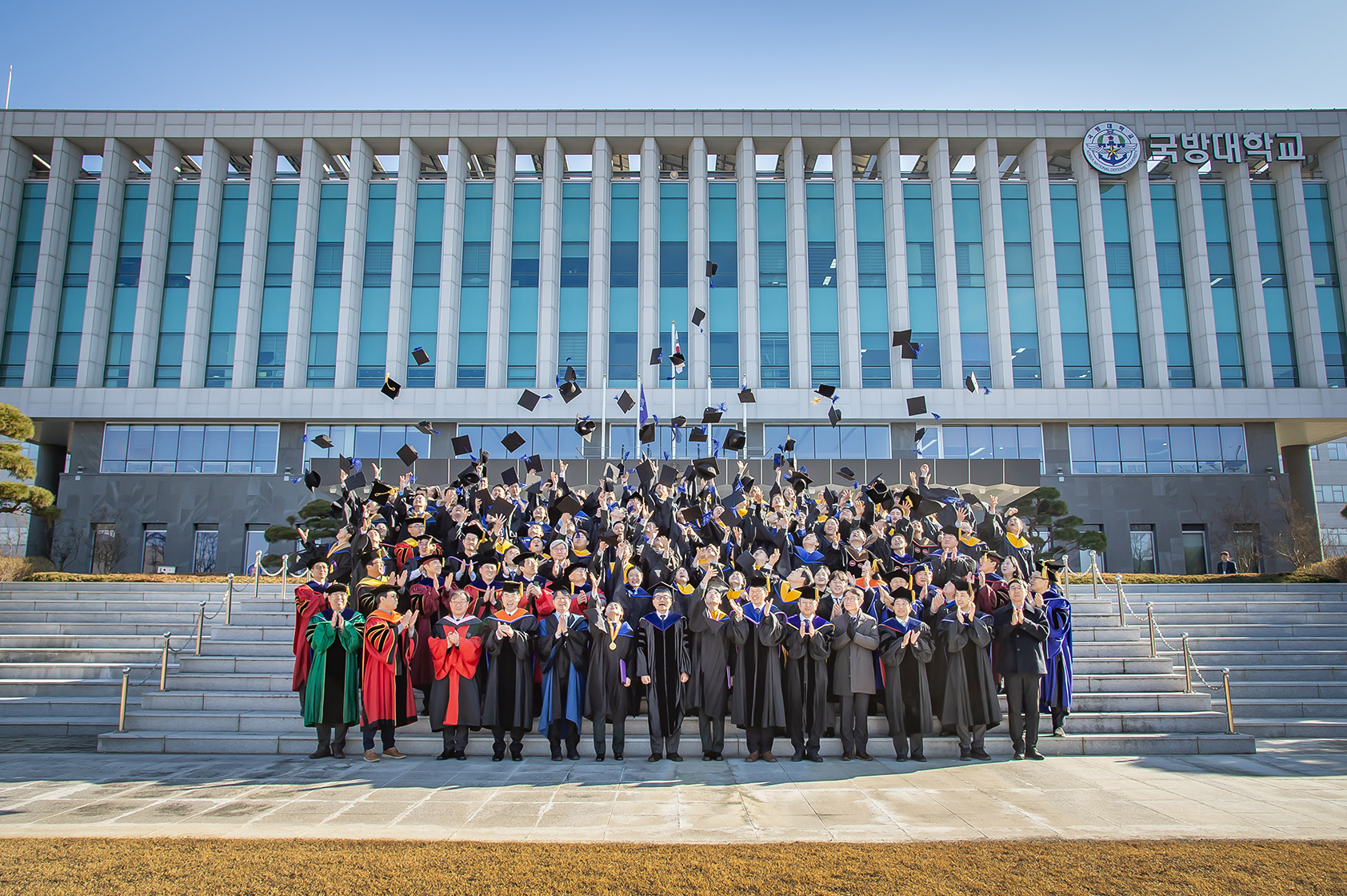 학위과정 학위수여식이 끝나고 국방대학교 앞에서 학위를 수여받은 학생들이 단체로 학사모를 하늘위로 던지고 있는 사진이다.